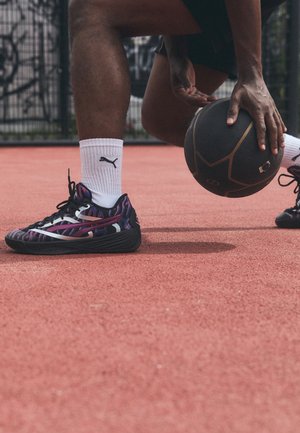 Personne dribblant un ballon de basket noir sur un terrain rouge extérieur, portant des baskets noires et violettes et des chaussettes blanches Puma.
