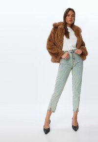 Ruskea tekoturkistakki, valkoinen t-paita, vaaleansiniset korkeat farkut, joissa on sivuleikkaukset, ja mustat korkokengät. Malli seisoo.