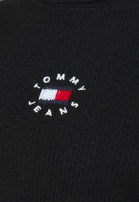 Czarny sweter z dzianiny z okrągłym logo, na którym u góry znajduje się napis "TOMMY", a na dole "JEANS", ozdobiony czerwonymi, białymi i niebieskimi poziomymi pasami.