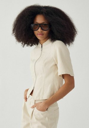 Kvinde med stort krøllet hår iført store mørke solbriller, en beige kortærmet lynlåsbluse og matchende bukser med hænderne i lommerne.