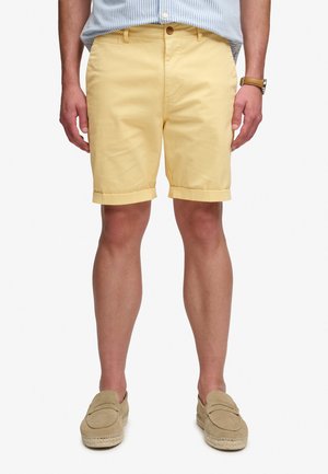 Mand iført lysegule shorts med opbuk, en blåstribet skjorte stoppet ned i, lysebrune slip-on sko og en brun ur på venstre håndled.