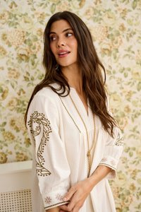 Kvinde med langt brunt hår iført en hvid kjole med grøn broderi på ærmerne og en guldkæde, stående foran tapet med blomster.