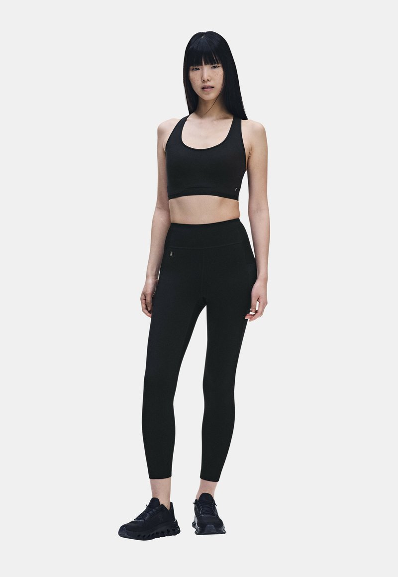 Femme debout portant un soutien-gorge de sport noir, des leggings taille haute et des chaussures de course noires sur un fond clair uni.