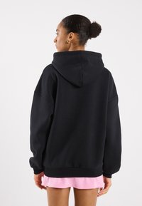 Felpa nera oversize con cappuccio, tessuto morbido, spalle scese e polsini a coste, indossata sopra un vestito mini rosa.