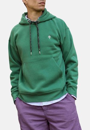 Homme portant un sweat à capuche vert avec un logo d'arbre et un pantalon violet, mains dans les poches, regardant sur le côté.