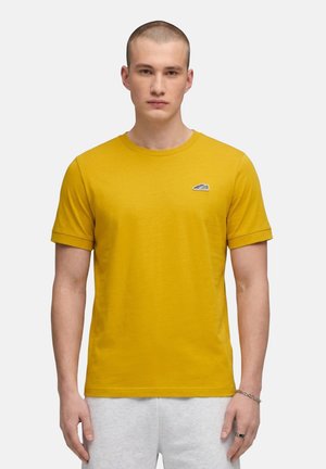 Giovane uomo con una maglietta giallo senape con un piccolo logo di sneaker sul petto e pantaloni grigio chiaro, in piedi rivolto in avanti.