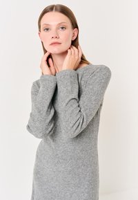 Pull en maille gris avec une coupe décontractée et une texture côtelée. Il présente de longues manches légèrement bouffantes et un col large. Design minimaliste.