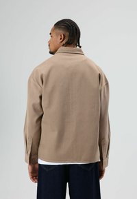 Veste-chemise en coton marron avec col, poignets boutonnés et coupe décontractée. Présente une texture lisse et un design minimaliste.