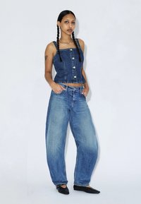 Femme aux cheveux tressés portant un top en denim sans manches boutonné, un jean large en denim et des ballerines noires pointues, debout sur un fond blanc.