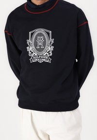 Sudadera de cuello redondo en color navy con costuras rojas, que presenta un logotipo bordado en blanco y una textura de tela diferenciada. Puños y dobladillo de canalé, con un corte relajado.