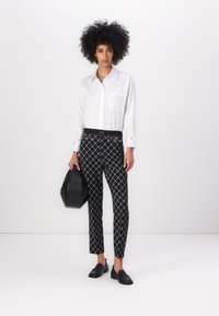 White button-up shirt with pocket; black patterned trousers featuring gold accents; black loafers; holding a black structured handbag.