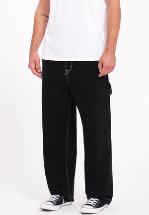Black cargo pants made of denim, featuring white stitching, a straight cut, and a side pocket. Paired with black canvas sneakers.