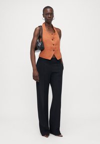 Oranje cropped vest met knopen, gedragen met zwarte wijde broek en puntige leopard print hakken. Houdt een kleine zwarte tas.