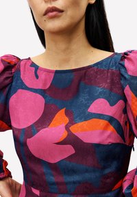 Eine Frau trägt ein dunkelblaues Kleid mit großen abstrakten rosa und orangefarbenen Blumenmustern, gepufften Ärmeln und einem runden Ausschnitt.