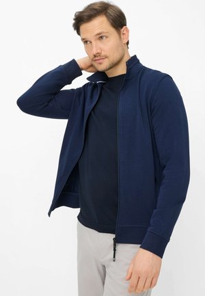 Man in marineblauw jack met rits en een zwart shirt, met lichtgrijze broek, die de achterkant van zijn nek aanraakt en opzij kijkt.