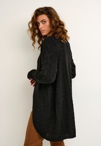 Zwarte gebreide cardigan met een lange, losse pasvorm, voorzien van een gestructureerd golvend patroon en doorgezakte schouders, gecombineerd met een bruine broek.