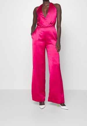 Kobieta ubrana w jaskrawy różowy satynowy kombinezon na szyję z szerokimi nogawkami i białe buty na szpic na jednolitym tle.