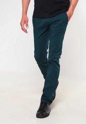 Man loopt en draagt strak zittende donker groene broek, zwart shirt en zwarte leren sneakers, linkerhand in de zak.