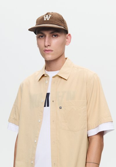 Chemise beige à manches courtes avec un motif à carreaux et une poche poitrine, superposée sur un t-shirt blanc, coiffée d'une casquette marron portant un logo.