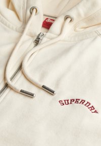 Haut zippé beige en tissu doux. Il présente un logo "SUPERDRY" brodé en rouge et des embouts argentés sur les cordons de serrage.