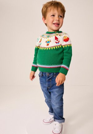 Grüner Strickpullover mit weißem Schulterbereich und bunten Weihnachtsmotiven. Kombiniert mit blauen Jeans und weißen Turnschuhen.