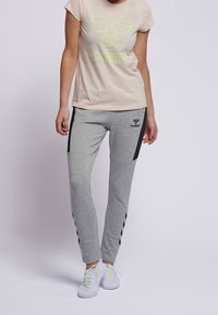 Camiseta beige claro con logo verde neón, combinada con pantalones de chándal grises que tienen paneles laterales negros y un logo. Zapatillas blancas con acentos neón.