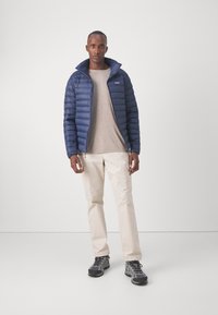 Tengerészkék puff jacket magas gallérral, texturált anyaggal, cipzáros elülső résszel, bézs környakas pólóval, világos bézs nadrággal és szürke túracipővel.