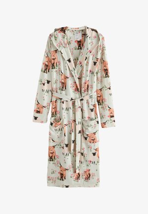 Femme portant une robe de chambre vert clair longueur mollet avec motifs de vaches et fleurs, debout pieds nus, mains sur les hanches, sur fond blanc.