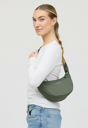 Vrouw met gevlochten haar, draagt een wit shirt met lange mouwen en spijkerbroek, draagt een groene crossbodytas over haar schouder, kijkt opzij.