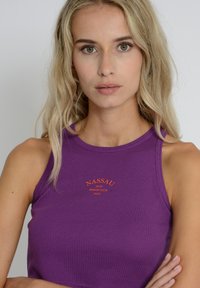Sleeveless purple ribbed tank top featuring an orange embroidered logo "NASSAU IBIZA BEACH CLUB 2008" on the chest area.