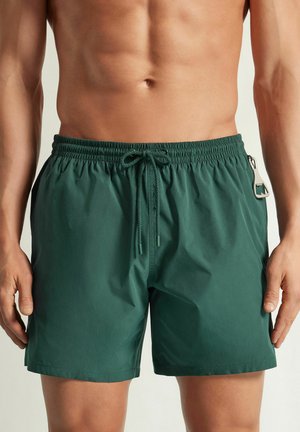 Mann trägt grüne Badeshorts mit elastischem Bund und Kordelzug, mit einem an der Seite angebrachten Flaschenöffner.
