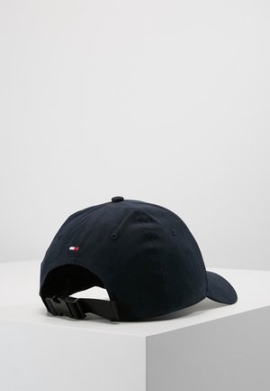 Czarna regulowana czapka z daszkiem z małym czerwono-biało-niebieskim prostokątnym logo z tyłu, spoczywająca na białej powierzchni na tle szarego tła.