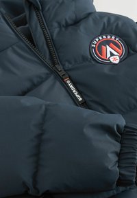 Námornícky pufrový kabát s prešívaným dizajnom, ktorý má čierny zips a okrúhlu vyšívanú nášivku na hrudi. Mäkkučkú textúru.