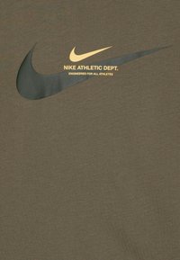 Olive spalvos Nike marškinėliai su dideliu juodu ir aukso spalvos Nike swoosh logotipu. Ant jų yra užrašas "NIKE ATHLETIC DEPT." ir "ENGINEERED FOR ALL ATHLETES."