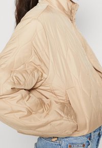 Beżowa pikowana kurtka bomberka z wysokim kołnierzem i zamkiem błyskawicznym. Posiada puchowe rękawy i subtelny połysk, wykonana z lekkiego materiału.