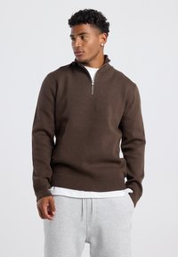 Brown sweater with a half-zip collar, ribbed cuffs and hem, made from soft fabric; worn over a white shirt and grey joggers.
