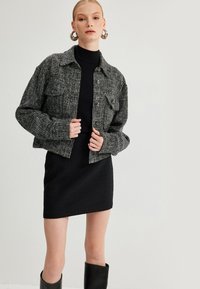 Grijze tweedjas met voorzakken, knoopsluiting en lange mouwen; gecombineerd met een zwart getextureerde jurk en kniehoge zwarte laarzen.