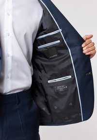 Marineblauer Blazer mit strukturiertem Muster, satinierter Fütterung, hellblauen Akzenten, inneren Taschen und sichtbaren Markenetiketten. Weißes Hemd teilweise sichtbar.
