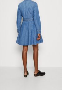 Denim-mekko pitkine hihoineen, vyötäröllä vyö, levenevä helma ja napit manseteissa. Yhdistetty mustiin puukengän, joissa on kultaiset niitit.