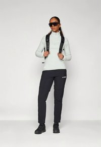 Világosszürke, hosszú ujjú, cipzáras felső, fekete nadrág zsebekkel, fekete túracipő és napszemüveg. Hátizsák pántjai láthatóak a vállakon.