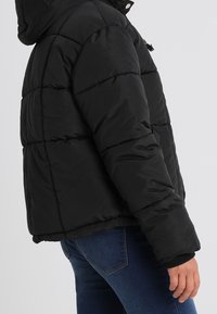 Person iförd en svart vadderad jacka och blå jeans, visad från sidan med armen avslappnat längs sidan.