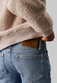 Felpa beige in pile con texture e polsini a costine, indossata sopra jeans azzurri chiari con una toppa in pelle marrone con logo sul retro.