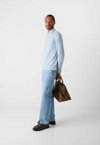 Mann iført lys blå langermet polo, lys blå jeans, mørkebrune sko, holder en brun skinnveske foran en enkel hvit bakgrunn.