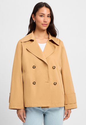 Beige double-breasted jacket with a wide collar, large buttons, and straight-cut sleeves. Worn over a white top and casual denim.
