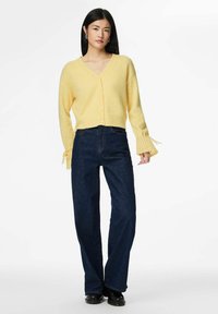 Żółty sweter na drutach z dekoltem w serek i zapięciem na guziki, z teksturowanymi bufkami na rękawach. Połączony z ciemnoniebieskimi, wysokimi spodniami jeansowymi.