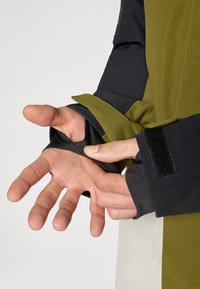 Texturovaná látka s dlhým rukávom v čiernej a olivovo zelenej farbe, so otvorom na palec a nastaviteľnými manžetami s suchým zipsom.