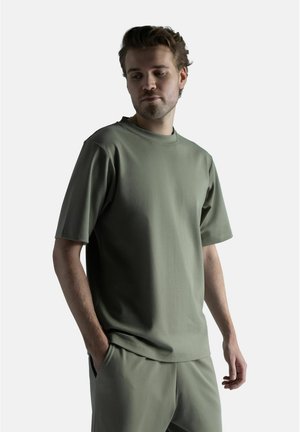 Nuori mies, jolla on yllään yhteensopiva oliivinvihreä lyhythihainen paita ja housut, seisoo valkoista taustaa vasten käsi taskussa.