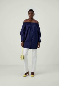 Mulher vestindo uma blusa azul marinho sem alças, calças brancas, bolsa amarela e sapatos de salto amarelos, de pé contra um fundo simples.