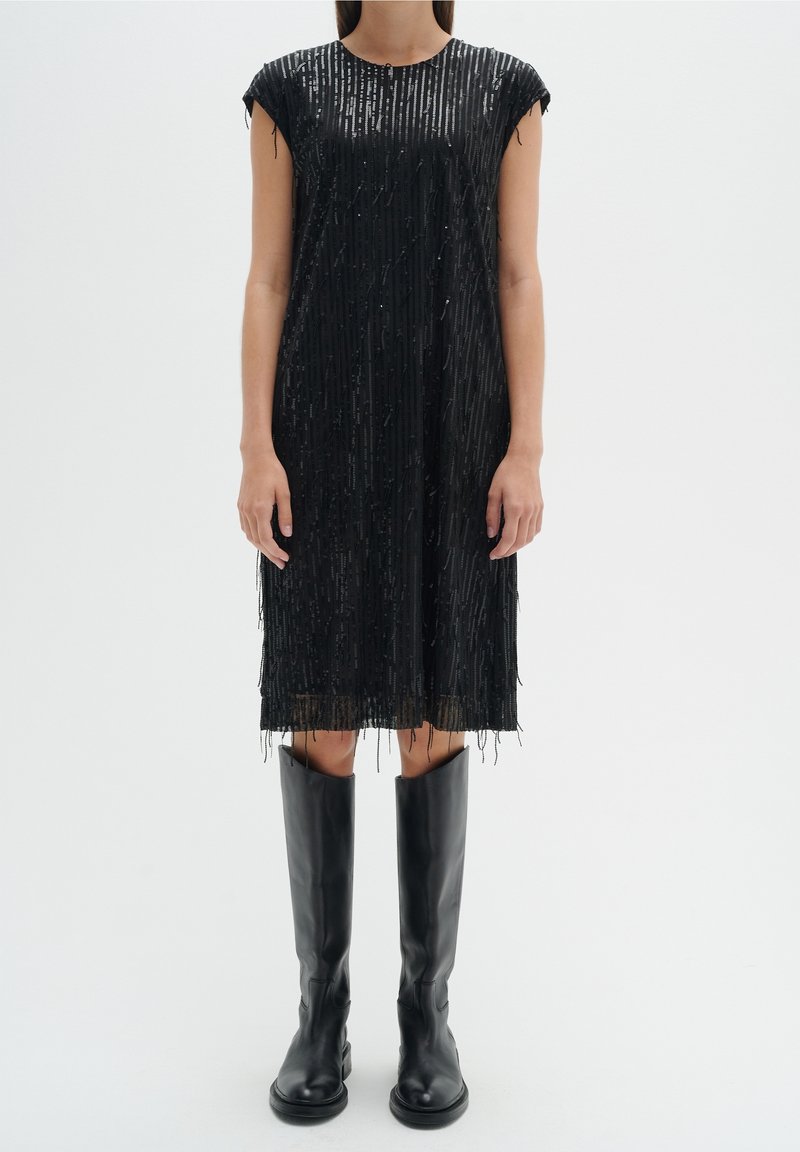 Černé šaty s vertikálními korálky, rovného střihu a s třásněmi na spodním lemu. Nosené s černými kozačkami nad kolena.