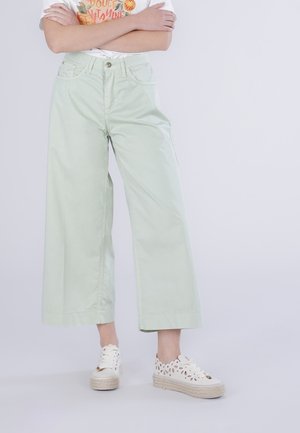 Person, der står med armene over kors iført lysegrønne vidde bukser, en hvid t-shirt med grafik og hvide plateau-sneakers med udskæringsdetaljer.